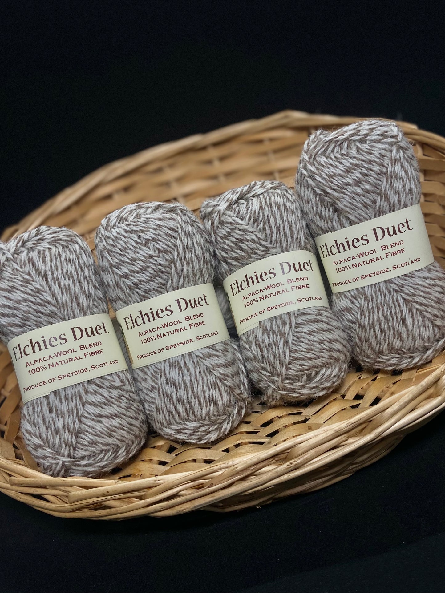 Four skeins of "Elchies Duet" wool displayed in a woven basket, in the shade "Light Marled". Packaging Transcription: "Elchies Duet. Alpaca-Wool Blend. 100% Natural Fibre. Produce of Speyside, Scotland".
