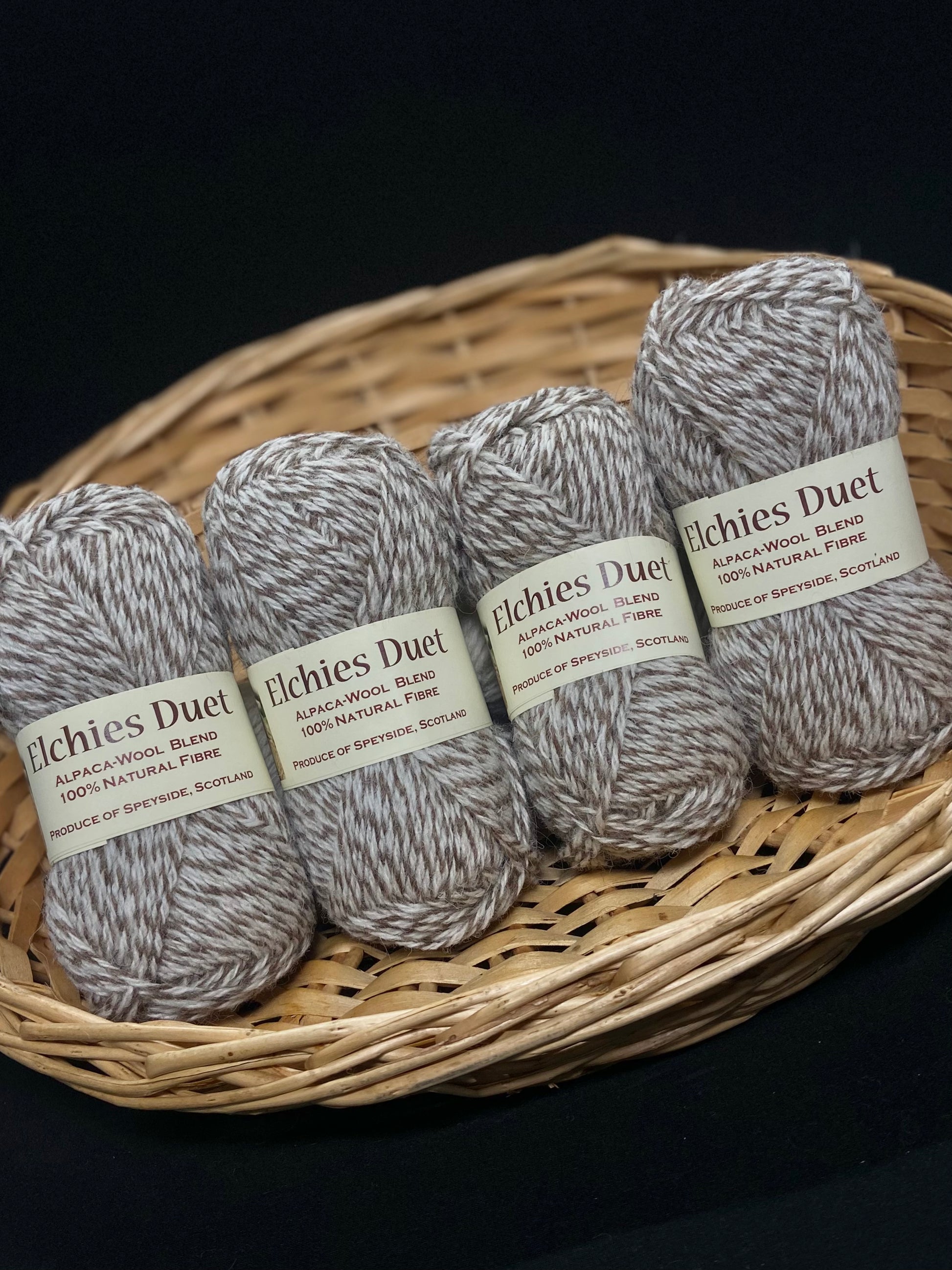 Four skeins of "Elchies Duet" wool displayed in a woven basket, in the shade "Light Marled". Packaging Transcription: "Elchies Duet. Alpaca-Wool Blend. 100% Natural Fibre. Produce of Speyside, Scotland".