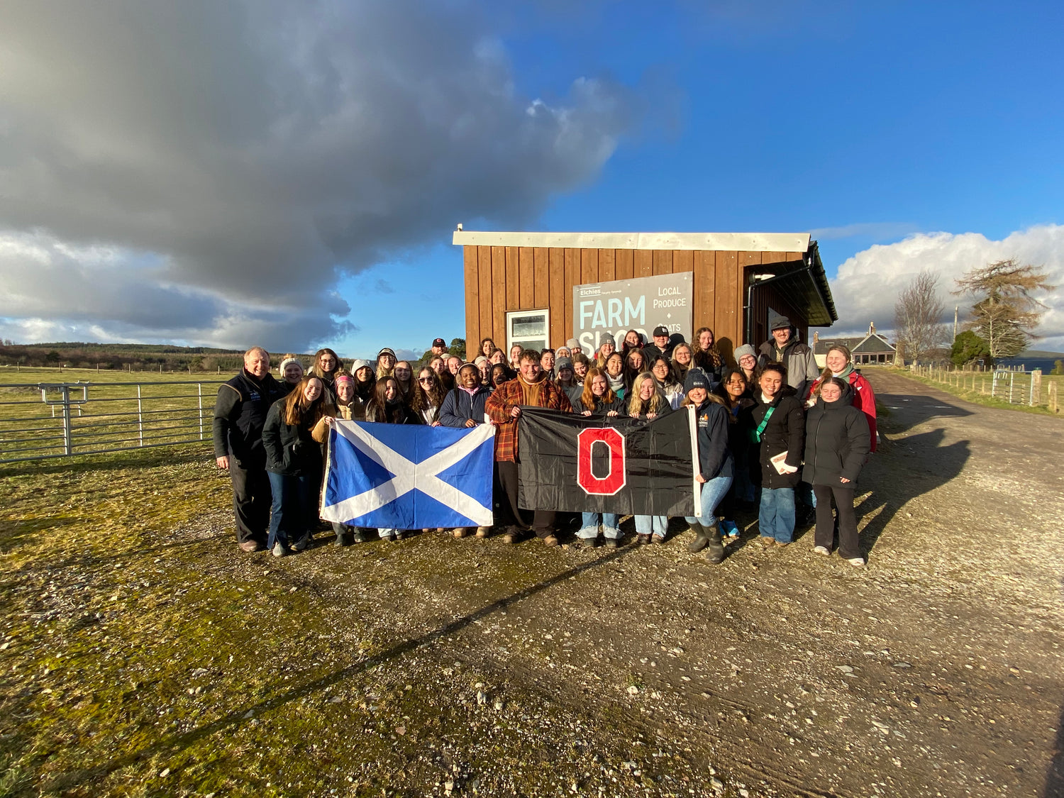 A photo of a large tour group visiting Elchies Farm, holding up a Scottish flag, and a black flag with a large, red 'O' in the middle.